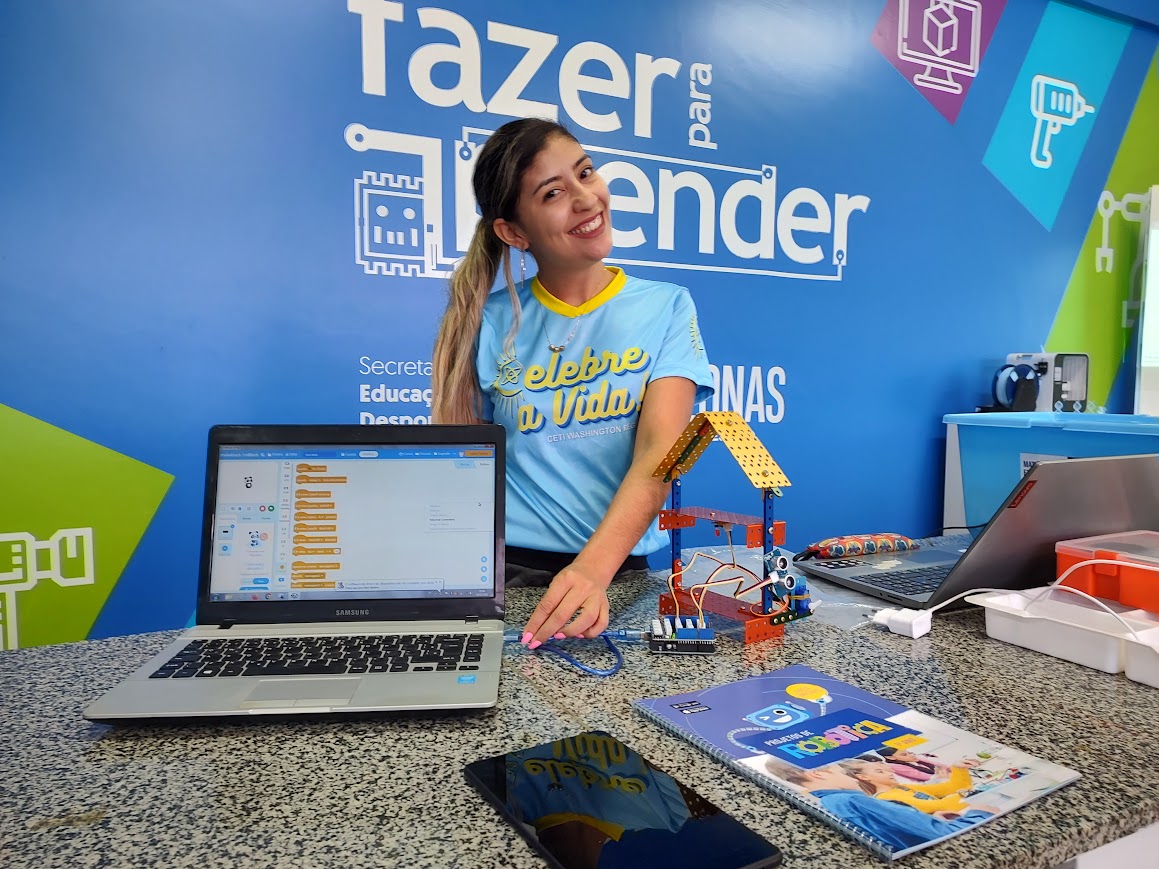 Educadora em ambiente de tecnologia educacional, ao lado de um notebook com programação visual aberta e um projeto de robótica montado sobre a mesa, representando aprendizagem prática em EdTech e pensamento computacional.